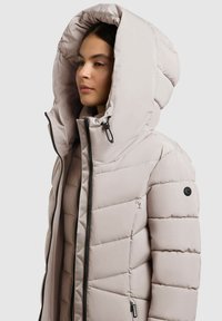 Svetlo bež puhovka s velikim kapuljačom, z sprednjim zadrgo, kvadratnim dizajnom in črnimi kovinskimi dodatki.