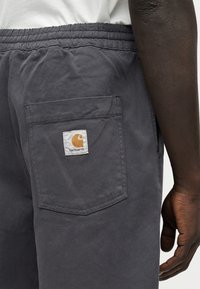 Άτομο με σκούρο δέρμα που φοράει γκρι παντελόνια Carhartt με ελαστική μέση και πίσω τσέπη με ετικέτα λογότυπου Carhartt.