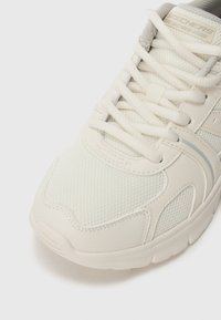 Baskets blanches Skechers Bobs avec des superpositions en mailles et en synthétique, des coutures détaillées et une semelle rembourrée sur fond blanc.