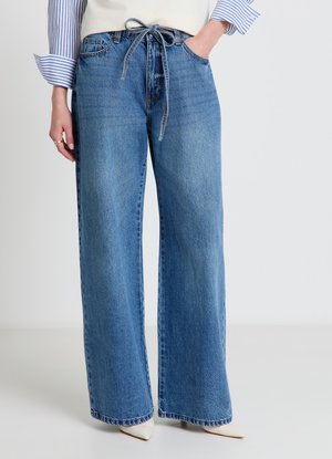 Frau trägt hoch taillierte, weit geschnittene blaue Jeans mit einem Denim-Gürtel, Hände in den Taschen, weiße spitze Schuhe und ein gestreiftes Hemd mit hochgekrempelten Ärmeln.