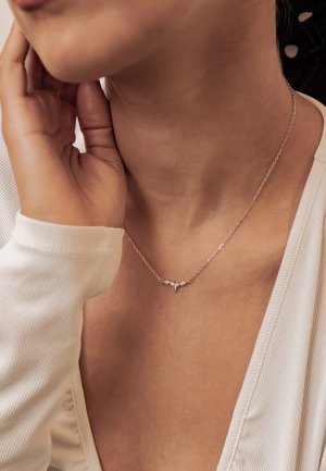 Frau trägt zarte silberne Halskette mit kleinen blattförmigen Anhängern, berührt ihr Gesicht, gekleidet in weißen, gerippten Oberteil.