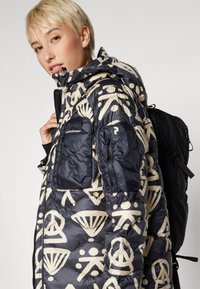 Casaco acolchoado em azul-marinho com padrões gráficos em creme. Apresenta um bolso com fecho no peito, capuz ajustável e uma textura resistente.