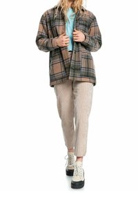 Chaqueta de franela de cuadros marrón, oversize, con acentos en verde y azul, combinada con una camiseta de color azul claro y pantalones de pana beige. Zapatillas negras de plataforma.