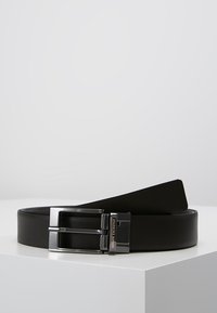 Ceinture en cuir noir avec une boucle en métal argenté à double ardillon, présentant un design épuré et une texture lisse. Logo de la marque gravé sur la boucle.