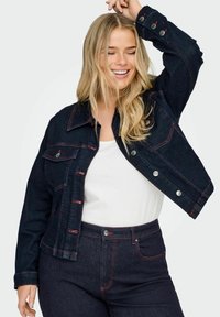 Leende kvinna med långt blont hår som bär en mörk denimjacka och byxor, vit topp, lyfter ena armen framför en neutral bakgrund.