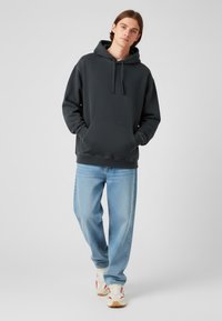 Donkergrijze hoodie met een buidelzak, verstelbare trekkoorden en een comfortabele pasvorm, gecombineerd met lichtblauwe jeans en witte sneakers.