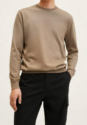 Homme portant un pull beige à manches longues et col rond, ainsi qu'un pantalon noir avec poches, debout avec une main dans la poche.