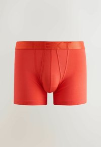 Boxeri bărbătești portocalii strălucitori, cu deschidere cusută și talie inscripționată cu "NEXT", pe un fundal simplu și deschis.