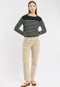 Frau steht mit hinter dem Rücken verschränkten Händen, trägt ein schwarz-weiß gestreiftes Langarmoberteil, beige Hosen und braune flache Schuhe vor einem weißen Hintergrund.