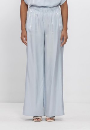 Vrouw die lichtblauwe satijnen wijde broek met elastische tailleband en witte sandalen met open tenen draagt, staande tegen een effen achtergrond.