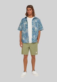 Camicia azzurra a maniche corte con un motivo a spirale bianco e verde, abbinata a pantaloni corti verde oliva e sneakers bianche con dettagli neri.