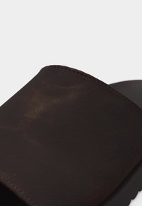 Κοντινό πλάνο ιμάντα καφέ σανδαλιού τύπου slide με ορατή ραφή και ματ, ελαφρώς υφή επιφάνεια.