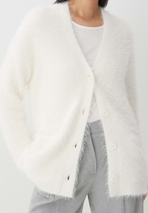 Femme portant un cardigan blanc duveteux boutonné par-dessus un haut blanc avec un pantalon gris clair, debout avec une main dans la poche.