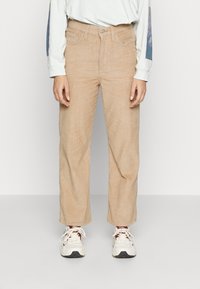 Pantalons en velours côtelé beige avec un design à jambes larges, dotés de poches avant et d'une fermeture à bouton. Portés avec une chemise à manches longues de couleur claire et des baskets.