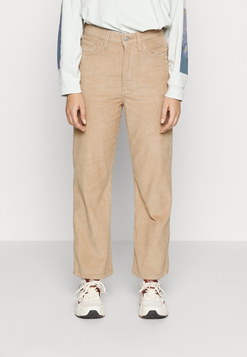 Pantalons en velours côtelé beige avec un design à jambes larges, dotés de poches avant et d'une fermeture à bouton. Portés avec une chemise à manches longues de couleur claire et des baskets.