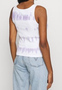 Top de tirantes de algodón blanco con patrón de tie-dye púrpura, diseño ajustado y tirantes anchos. La vista trasera muestra el dobladillo que termina a la altura de la cintura.