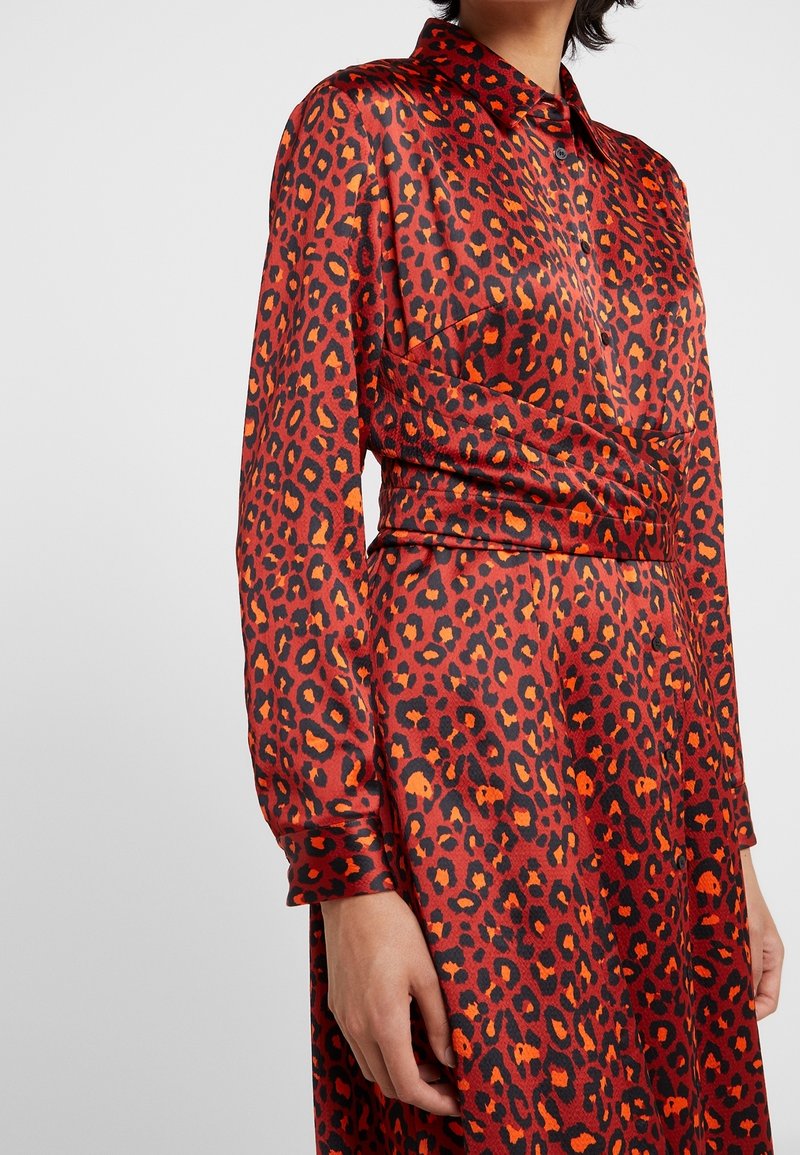 Femme portant un chemisier à manches longues rouge et orange imprimé léopard, avec une large ceinture assortie à la taille et une jupe.