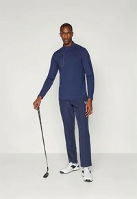 Marineblå, langermet treningstopp med halv glidelås, kombinert med matchende bukser. Modellen holder en golfklubbe og har på seg hvite treningssko.