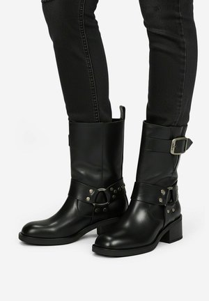 Stivali neri in pelle fino al polpaccio con fibbia d'argento e cinturino borchiato, indossati con pantaloni neri aderenti, su sfondo bianco.