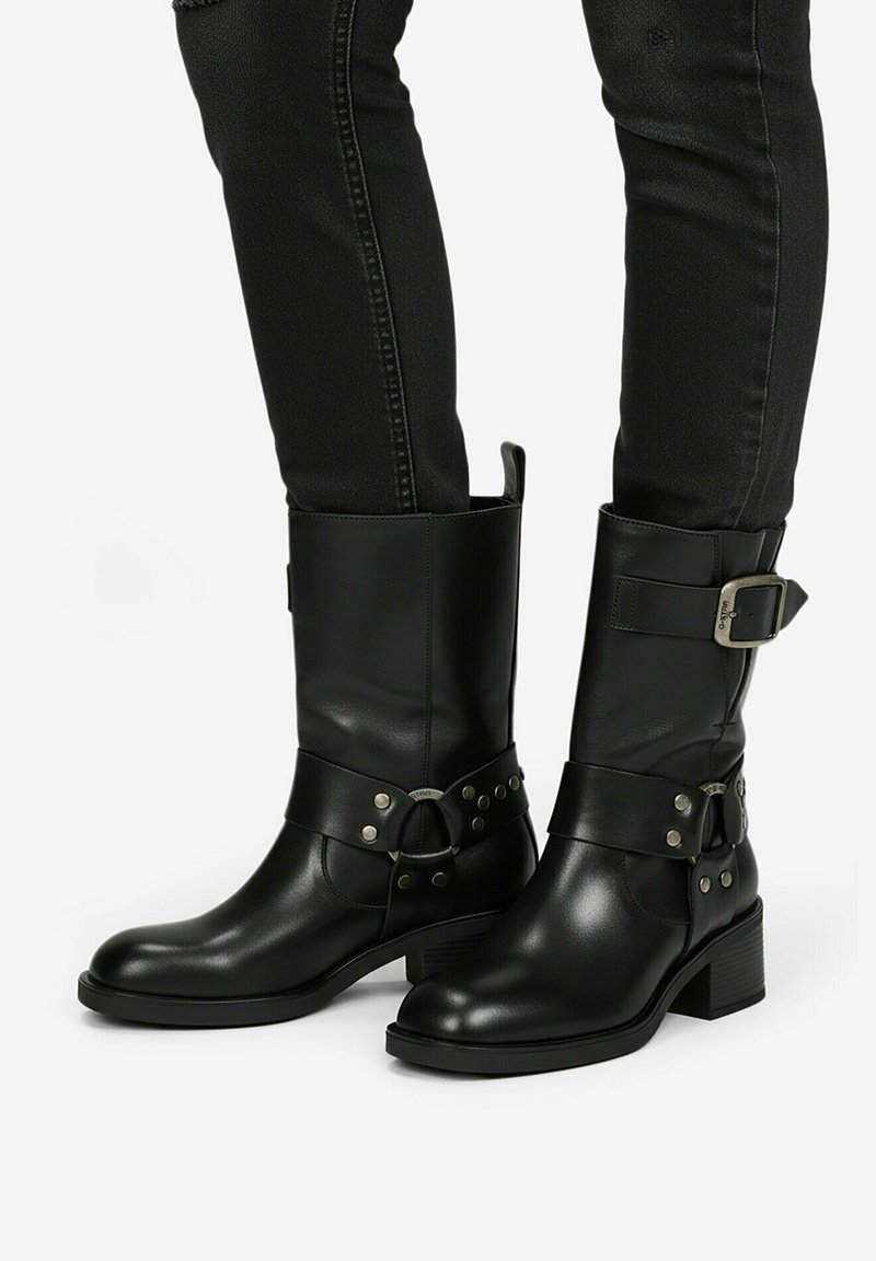 Botas negras de cuero hasta la mitad de la pantorrilla con hebilla plateada y correa tachonada, usadas con pantalones negros ajustados, sobre un fondo blanco.