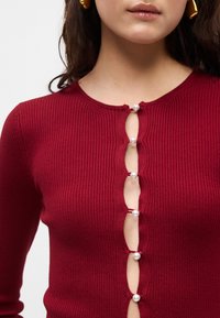 Ribota bordo megztinis su apvalia kaklo linija ir mygtukais priekyje, puikuojantis perlo tipo mygtukais ir banguotais iškirpimais.