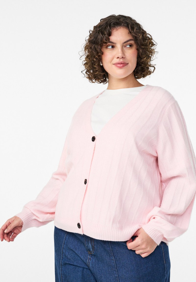 Light pink knitted cardigan with a v-neck, ribbed texture, and black buttons. Features long sleeves and relaxed fit. Worn over a white top.