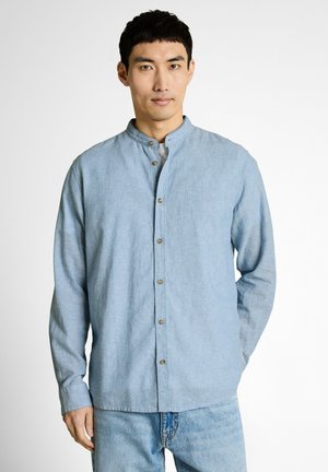 Mężczyzna w jasnoniebieskiej koszuli na guziki z kołnierzem mandarynkowym i niebieskich dżinsach, stojący na tle jednolitego białego tła.