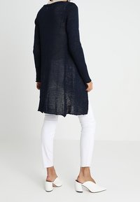 Marinblå stickad lång kofta med en lös passform, öppna sidor och ett texturerat mönster, bärs över vita cropped jeans och vita mules.