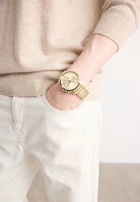Montre-bracelet en or avec un cadran texturé, un bracelet en métal et des sous-cadrans, portée sur un pull et un pantalon de couleur claire. Design minimaliste et fonctionnel.