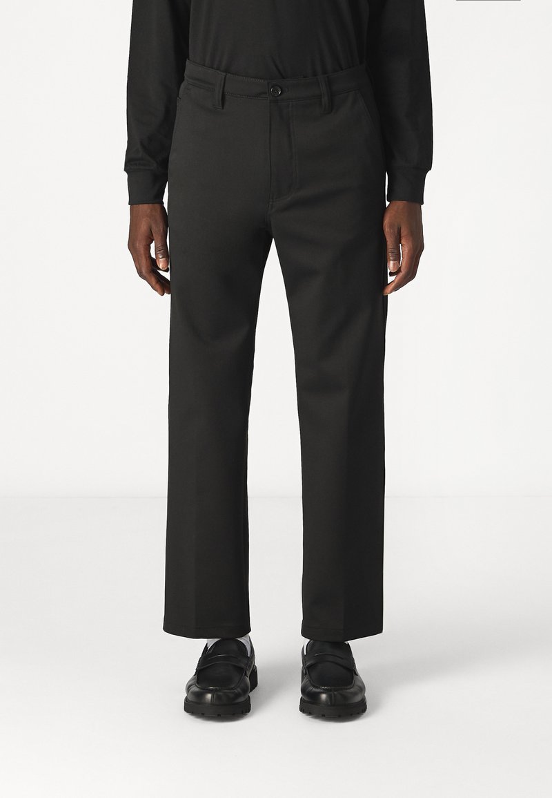 Pantalon noir à jambe droite en tissu lisse, doté d'une fermeture à bouton, de passants de ceinture et de poches discrètes ; porté avec des chaussures noires.
