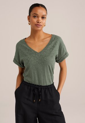 T-shirt vert olive à col en V avec des manches courtes retroussées, présentant un motif léopard texturé. Associé à un pantalon noir à taille élastique.