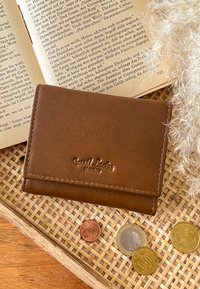 Rudas odinis piniginė su lygiu paviršiumi ir iškilniu logotipu. Padėta ant pintos lėkštės kartu su monetomis ir atvira knyga.