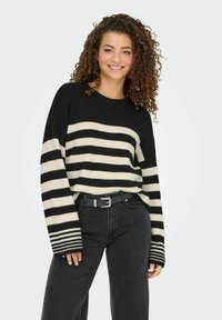 Zwart en crèmekleurig gestreepte trui met een geribbelde textuur, ronde halslijn en een licht oversized pasvorm, gecombineerd met hoge zwarte jeans.
