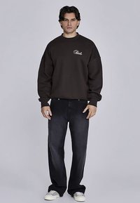 Donkerbruine sweater met ronde hals en geborduurd logo, gecombineerd met losse zwarte jeans en witte sneakers, in een frontale houding.