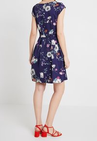 Abito floreale navy con maniche a cappuccio, vita aderente, gonna a ruota e motivi di fiori colorati. Indossato con sandali rossi con cinturini.