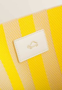 Tissu rayé jaune et beige avec une étiquette rectangulaire blanche arborant un design de tortue dorée. Le matériau semble texturé et tissé.