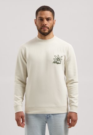 Man draagt een crèmekleurige trui met lange mouwen, met een donkergroene bloemafbeelding en tekst op de linkerborst, gecombineerd met lichtblauwe spijkerbroek.