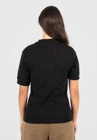 Donna con lunghi capelli castani che indossa una maglietta nera a maniche corte e pantaloni marroni, in piedi con le spalle rivolte verso la telecamera su uno sfondo neutro.