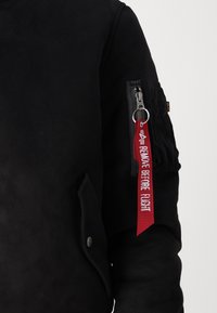 Giacca bomber nera in tessuto testurizzato, con una tasca laterale con zip e un'etichetta rossa "RIMUOVERE PRIMA DEL VOLO" attaccata. Presenta una chiusura con bottoni a pressione.