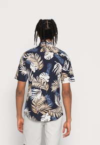 Camicetta a maniche corte con uno sfondo blu navy e grandi stampe tropicali di foglie e ananas bianche, beige e blu. Tessuto morbido, vestibilità comoda.