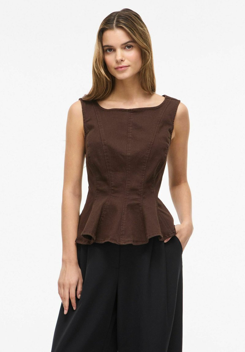 Femme portant un top péplum marron sans manches et un pantalon noir ample, debout avec une main dans la poche devant un fond blanc uni.
