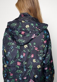 Femme aux longs cheveux blonds portant une veste de pluie sombre à motif floral avec une capuche, vue de dos sur un fond uni.