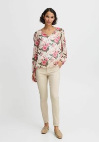Blumige Bluse mit rosa und weißen Rosenmustern, halbtransparentem Stoff und langen Ärmeln. Kombiniert mit beigen, engen Hosen und leichtem Schuhwerk.