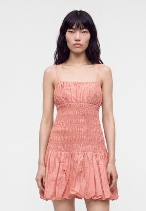 Jeune femme aux longs cheveux foncés portant une robe rose sans manches froncée avec bretelles spaghetti sur un fond uni.