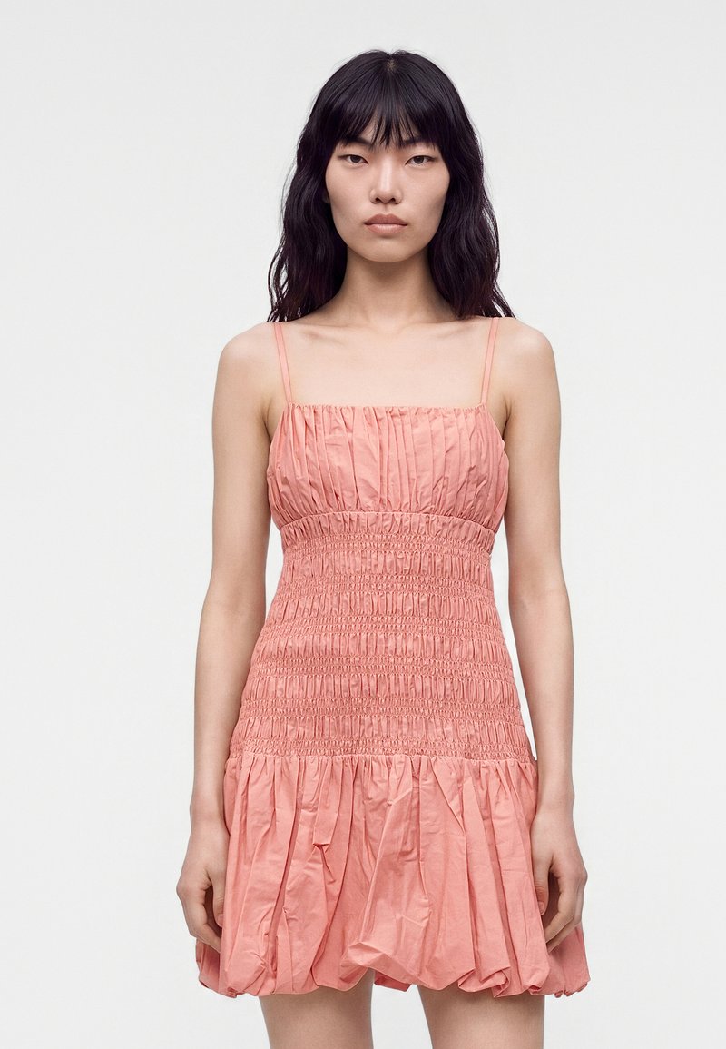 Jeune femme aux longs cheveux foncés portant une robe rose sans manches froncée avec bretelles spaghetti sur un fond uni.