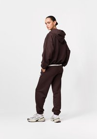 Brązowa bluza z kapturem i dopasowane spodnie dresowe wykonane z miękkiego materiału. Charakteryzują się luźnym krojem, przednią kieszenią i bocznymi paskami. Noszone ze sneakersami.