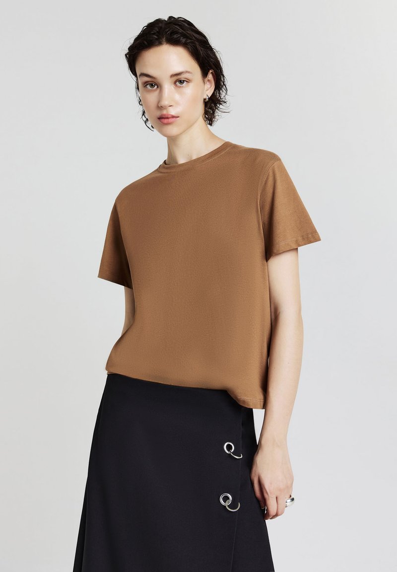 Giovane donna con capelli scuri e corti, vestita con una maglietta marrone a maniche corte e una gonna nera con dettagli in anello argentato, posando davanti a uno sfondo semplice.