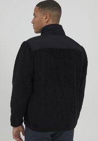 Svart fleecejakke med høy krage, laget av et teksturert, fluffy materiale med glatte nylon-skuldrer. Vist fra baksiden.