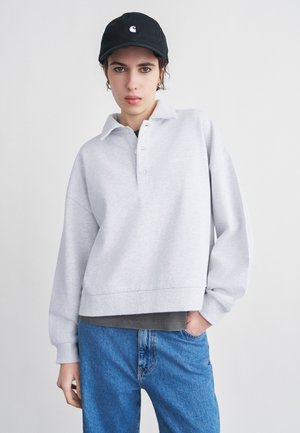 Donna che indossa un maglione grigio chiaro con bottoni, jeans blu e un cappello nero, in piedi davanti a uno sfondo bianco uniforme.