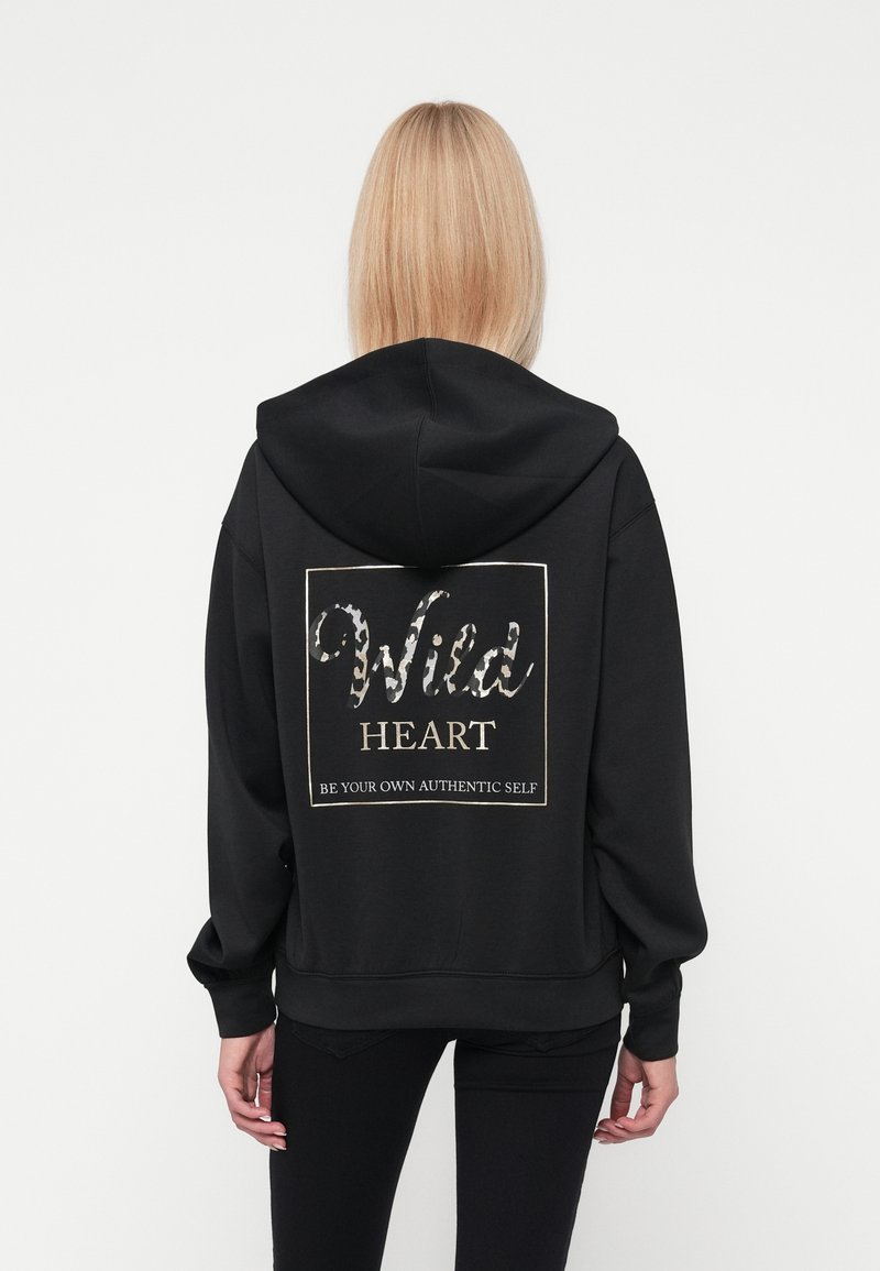 Fekete kapucnis pulóver, amelyen egy textúrázott grafika található, a hátsó részén "Wild HEART" és "BE YOUR OWN AUTHENTIC SELF" feliratok fehér és bézs színben.